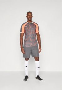 Šedé atletické tričko s oranžovými akcentmi a vzorom vĺn, doplnené o šedé šortky, biele ponožky a čierne športové topánky.