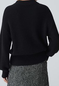 Zwarte gebreide trui met een geribde textuur, voorzien van een hoge kraag en raglanmouwen, gecombineerd met een gestreepte zwart-wit rok.