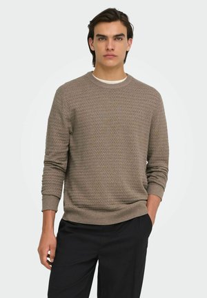 Jeune homme aux cheveux foncés portant un pull marron texturé à manches longues et un pantalon noir, debout avec une main dans la poche.