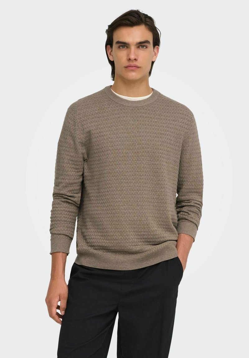 Jeune homme aux cheveux foncés portant un pull marron texturé à manches longues et un pantalon noir, debout avec une main dans la poche.