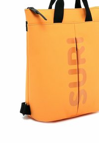 Pomarańczowa torba na ramię z czarnymi paskami, wykonana z gładkiego, trwałego materiału. Posiada centralny zamek błyskawiczny oraz duże, wyraźne logo „SURI” na przodzie.