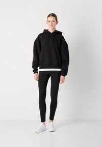 Black oversized hoodie with drawstrings, worn over a white strip. Paired with black leggings and white sneakers, set against a plain background.