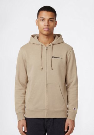 Młody mężczyzna ubrany w beżową bluzę Champion na zamek z kieszeniami z przodu i czarne spodnie, stojący na białym tle.