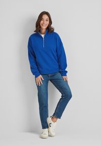 Blue sweatshirt with a quarter-zip collar, relaxed fit, worn with blue jeans and white sneakers, displayed against a neutral backdrop.