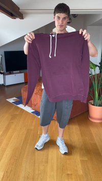 Bordo spalvota su kapuce, su raišteliais, priekiniais kišeniais ir įvarstytais rankogaliais. Modelis laiko ją prieš švelniai apšviestą interjero aplinką su medinių grindų danga.