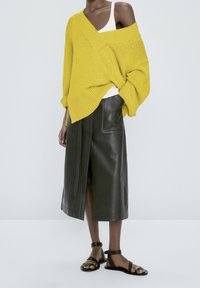 Femme portant un pull oversize jaune moutarde tombant sur une épaule, un débardeur blanc, une jupe midi en cuir noir fendue à l'avant et des sandales noires.