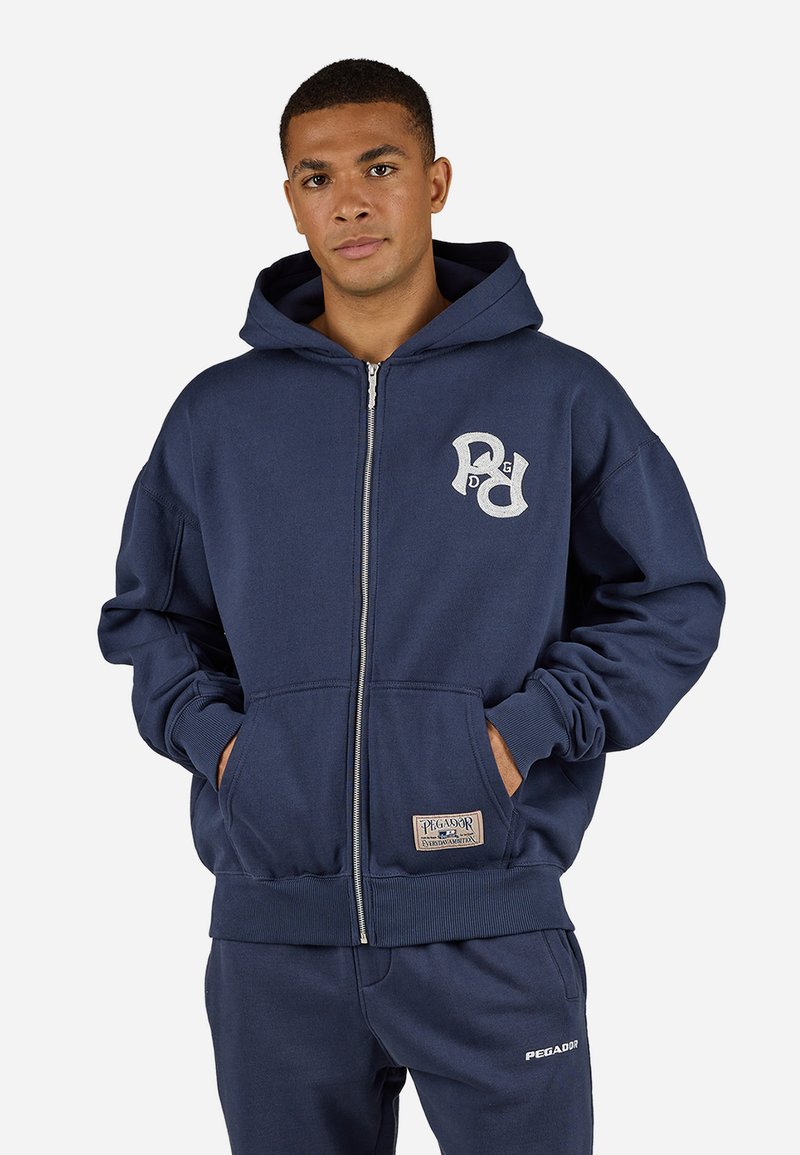 Navy blue zip-up hoodie with a front pocket, logo on the chest, ribbed cuffs, and a soft texture. Paired with matching pants.