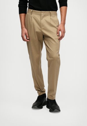 Pantalons taillés beige avec une texture lisse, devant plat et deux poches latérales, associés à des baskets noires de sport.