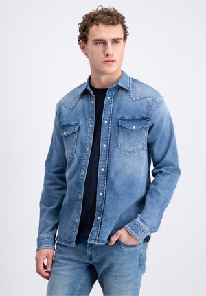 Jonge man met krullend haar, die een lichtblauw spijkerjack draagt over een zwart shirt en bijpassende blauwe jeans, staand met één hand in de zak.