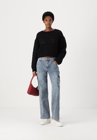 Svart strikket crop-genser med synlige sømmer, lyseblå vide cargo-jeans, hvite sneakers og en liten rød håndveske.