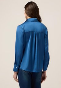 Blusa di raso blu con colletto, maniche lunghe e polsini con bottoni. Presenta una piega sul retro e una superficie liscia e lucida.