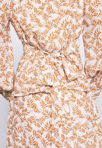 Bluse aus einem blumenmusterigen Stoff in hellbeige mit orangefarbenen Blattmotiven; sie hat eine Bundfalten mit Bindeband und geraffte Ärmeldetails.