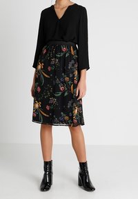 Haut noir avec décolleté en V et manches longues, associé à une jupe noire mi-longue ornée de motifs floraux colorés. Porté avec des bottines noires brillantes.