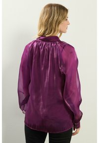 Blusa trasparente viola con yoke arricciato, maniche lunghe e voluminee, polsini con bottoni e una superficie liscia e lucida.