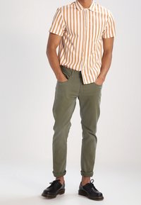 Muž v krátkém rukávu, s oranžovo-bílým pruhovaným tričkem, olivově zelenými kalhotami s ohrnutými nohavicemi a černými šněrovacími botami, s rukama v kapsách.