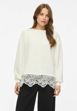 Un mannequin se tient devant un fond blanc, portant un haut crème à manches longues avec un ourlet en dentelle et un pantalon noir.