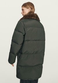 Woman wearing a long dark green puffer coat with a brown fur collar, viewed from the back against a plain background.