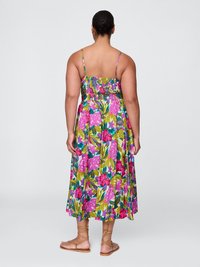Frau mit dunkelhaarigen Dutt, die ein ärmelloses, blumiges Midi-Kleid mit dünnen Trägern und beigen, schnürbaren Flachsandalen trägt, mit dem Rücken zur Kamera auf einem einfarbigen Hintergrund.