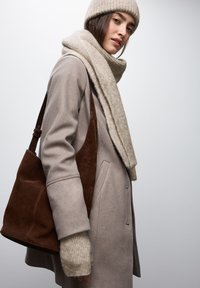 Cappotto di lana beige con colletto alto, abbinato a un cappello e una sciarpa beige a coste. Una borsa in suede marrone accentua l'outfit.