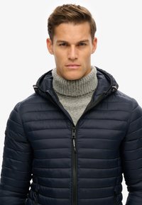 Marineblå pufferjakke med hætte, der har ribbet detaljer ved halsen. Stoffet er glat og skinnende, og fremhæver vandrette quiltede mønstre.