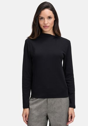 Black long-sleeve top with a crew neck and subtle gathered detail on the shoulder, paired with grey trousers. Smooth texture, casual fit.