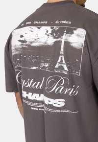 Šedé grafické tričko s velkým bílým potiskem Eiffelovy věže a textem "Crystal Paris CHAMPS" na zadní straně, krátké rukávy, volný střih.