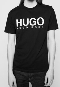 Camiseta de algodón negra con gran texto blanco "HUGO" y texto más pequeño "HUGO BOSS". Manga corta, escote redondo y un corte relajado.