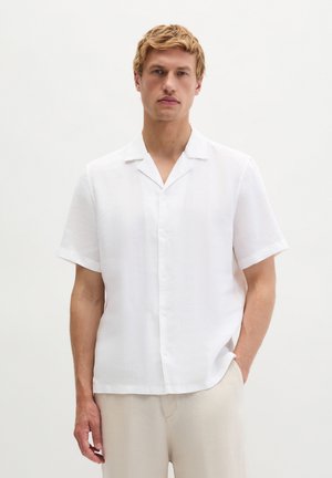 Blonder Mann trägt ein weißes kurzärmeliges Hemd mit Knöpfen und hellbeige Hose, steht vor einem einfarbigen hellen Hintergrund.