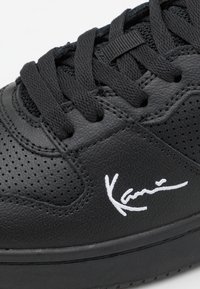 Fekete bőr sportcipő sima felülettel, légáteresztő perforációkkal és fehér hímzett logóval az oldalon. Fekete cipőfűzők és talp.