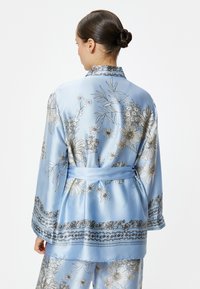 Túnica de satén azul con patrones florales en dorado y blanco, con cinturón de lazo y detalles en el dobladillo y las mangas.