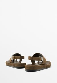 Sandales en suede marron avec une semelle noire plate, une sangle à boucle ajustable et un design ouvert à l'arrière pour un enfilage facile.