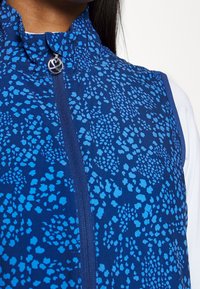 Blå mönstrad ärmlös väst med dragkedja fram över en vit långärmad topp, bärs av en person med mörkt hår synligt vid axeln.