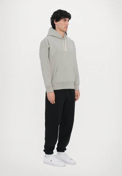 Hanorac gri deschis cu buzunar frontal și șnur, purtat cu pantaloni de trening negri și adidași albi. Design simplu și casual.