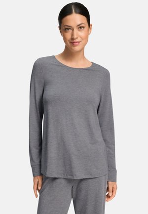 Donna con capelli scuri legati dietro che indossa una maglia grigia a maniche lunghe semplice e pantaloni grigi abbinati, in piedi e rivolta verso davanti.