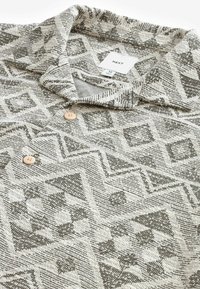 Camisa beige con un diseño geométrico y acentos en gris oscuro, cuello con solapa y dos botones beige en la tapeta.