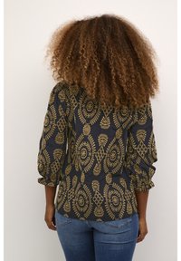 Svart blus med guld paisleymönster, texturerad tyg, trekvartslånga ärmar, samlat detaljer vid axlarna, ihop med blå jeans.