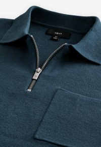 Mørkeblå zip-up trøje med struktureret strik, fold-over krave og frontlomme. Har en sølvfarvet lynlås og mærkelabel indeni.