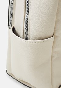 Beige texturerad fusk läderryggsäck med en sidoficka och metalliska dragkedjsaccent, som visar en minimalistisk och funktionell design.