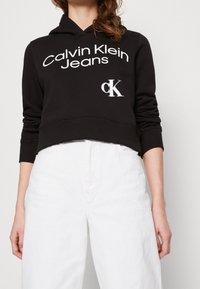 Sudadera negra recortada con un gran logotipo blanco de "Calvin Klein Jeans" y un emblema "ck". Cuenta con capucha con cordón y puños de canalé.