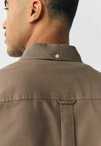 Bruine stoffen shirt met een gestructureerde kraag, drukknoopsluiting en een lusaccent onder de kraag. Zachte textuur en een nette afwerking.