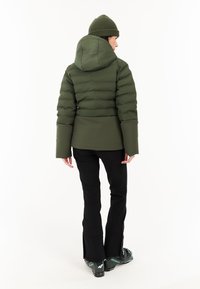 Gevoerde donkergroene jas met een capuchon, met horizontale naden en lange mouwen. Gepaarde met zwarte broek en donkere sneeuwlaarzen.