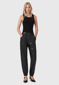 Jonge vrouw met blond haar, gekleed in een zwarte mouwloze top, zwarte leren joggingsbroeken en zwarte hoge hakken, staand met haar handen in de zakken.