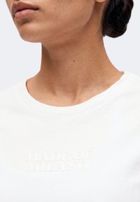 Gros plan sur le bas du visage et le cou d'une femme portant une chemise blanche avec le texte en relief « MADE OF MILANO » sur la poitrine.