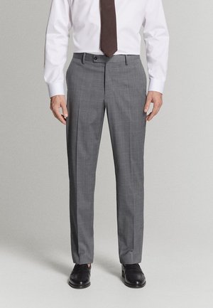 Homme portant un pantalon de costume gris à carreaux, une chemise blanche, une cravate marron et des chaussures de ville noires, debout devant un fond clair uni.