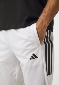 Pantalones deportivos blancos con diseño de tres rayas negras, que presentan un logo de Adidas, bolsillos laterales y una tela suave y ligera.