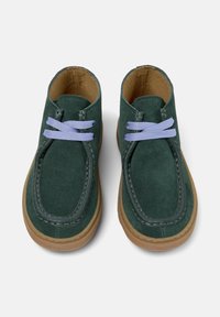 Zapatos de ante verde con suela oscura y cordones de un púrpura claro, vistos desde arriba, mostrando su forma redondeada y los detalles de costura.