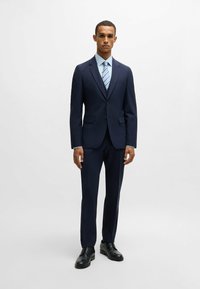 Marineblauer Anzug mit taillierter Passform, strukturiertem Stoff und Revers mit Einschnitten, kombiniert mit einem hellblauen Hemd und einer gestreiften Krawatte, ergänzt durch schwarze Schuhe.