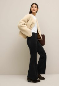 Cremfarbener, gefütterter, kurz geschnittener Mantel, weißes T-Shirt, weit geschnittene schwarze Hose und braune Knöchelstiefel. Strukturiertes Material mit dezenten Knopfdetails.