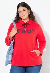 Punainen huppari, jossa on musta "TRUE" -painatus, naruhuppu, etutaskut ja pitkät hihat, yhdistettynä vaalean denimin laukkuun.