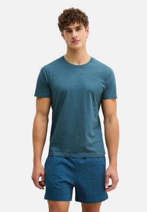 Man staand, gekleed in een turkoois T-shirt met korte mouwen en een ronde hals, en blauwe korte broek met patroon, tegen een witte achtergrond.